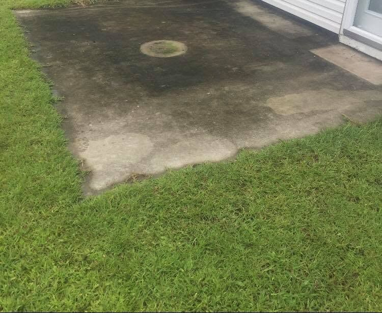 Weathered concrete patio with a small circular drain cover beside green grass and a house wall