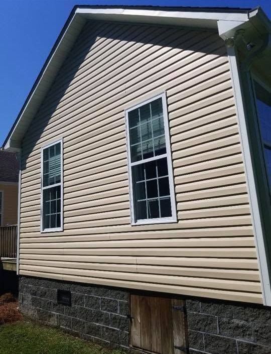 Beige vinyl-sided house exterior with two windows, stone foundation, and blue trim against a bright sky