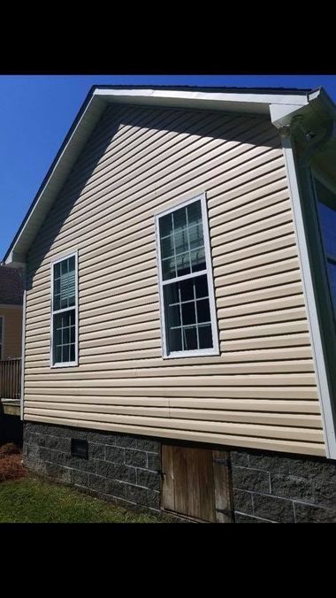 Beige vinyl-sided house exterior with two windows, stone foundation, and blue trim against a bright sky
