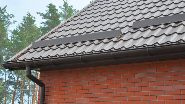Brick house roof with gray shingles and a gutter under cloudy sky