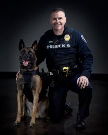 Police K-9 officer kneeling beside a German shepherd in dark uniform against a black background