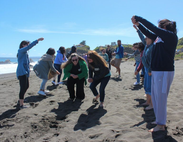 Gente jugando en una playa de arena negra con los brazos en alto. Día soleado, cielo azul y océano visible.