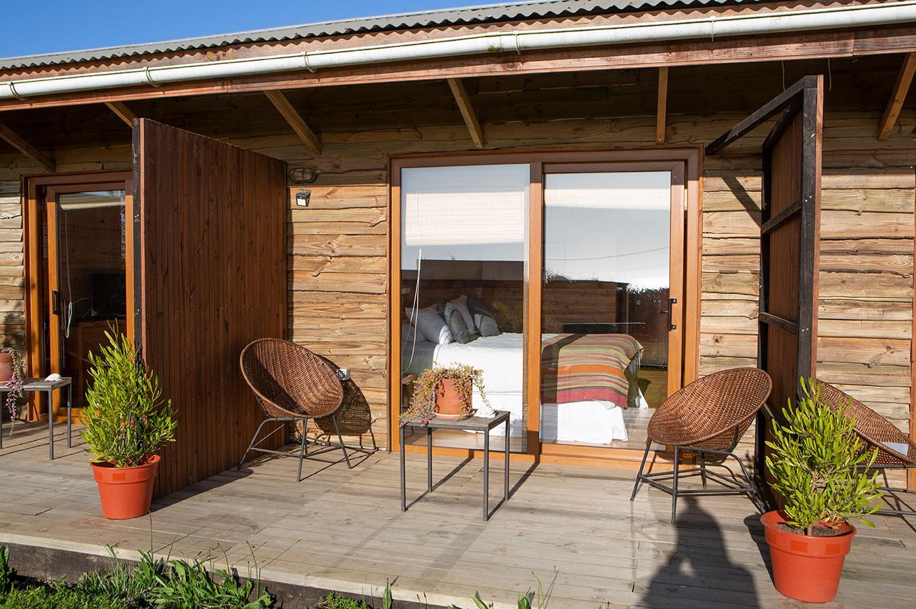 Exterior de cabaña de madera con patio, sillas de mimbre, plantas en macetas y puerta corrediza de vidrio.