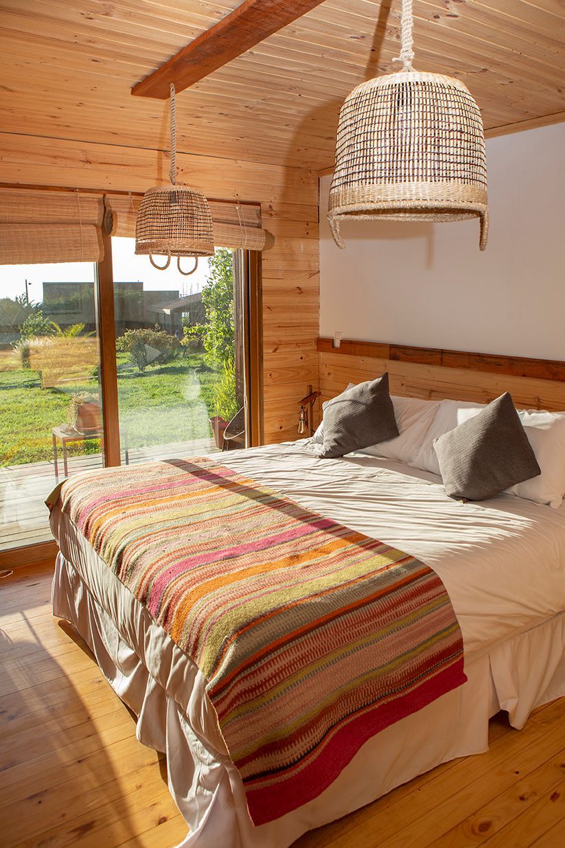 Dormitorio con paredes de madera, cama con manta de color y ventana que da a un patio verde.