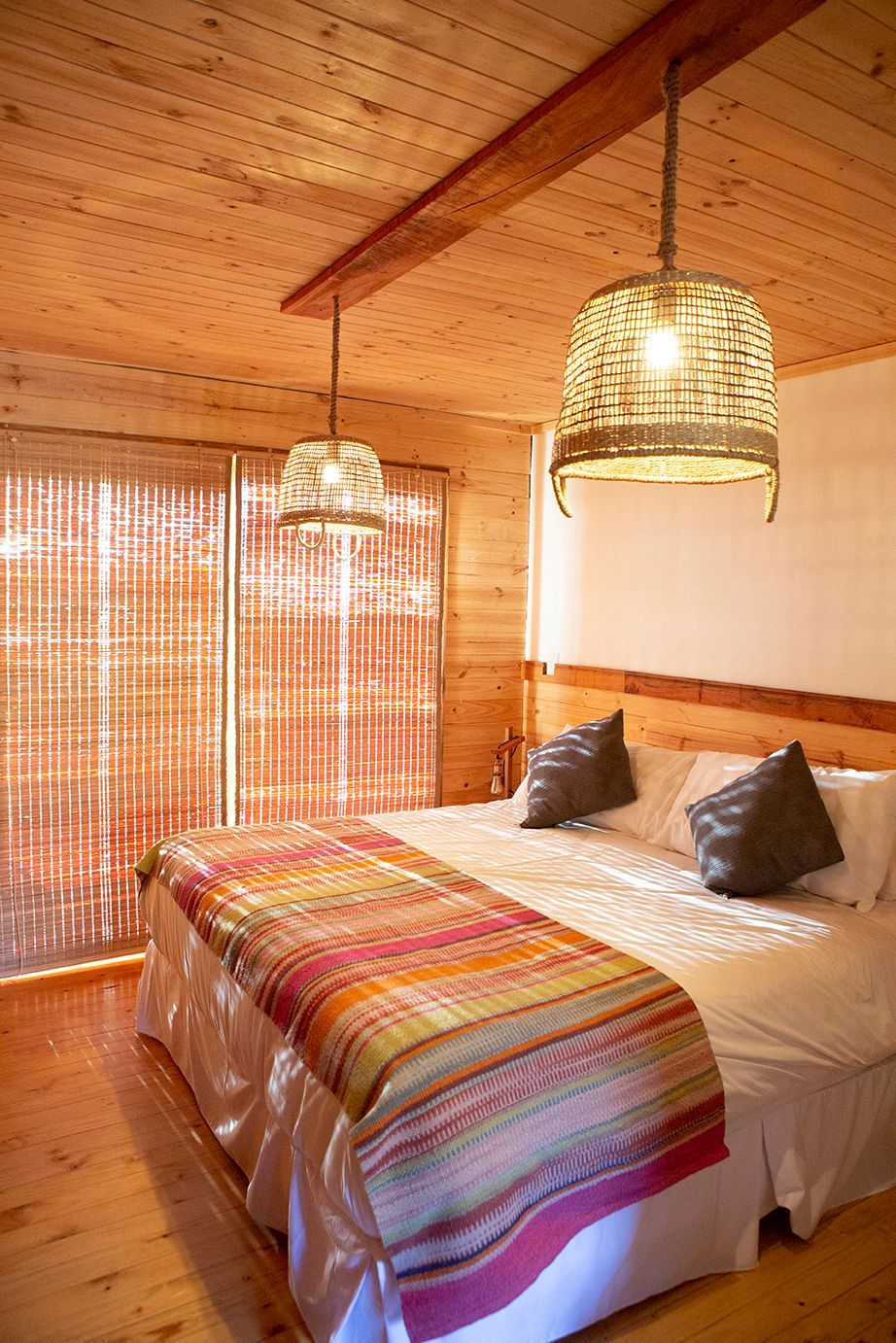 Dormitorio con paredes de madera, cama con manta de color, dos lámparas colgantes de mimbre y ventana con persianas.