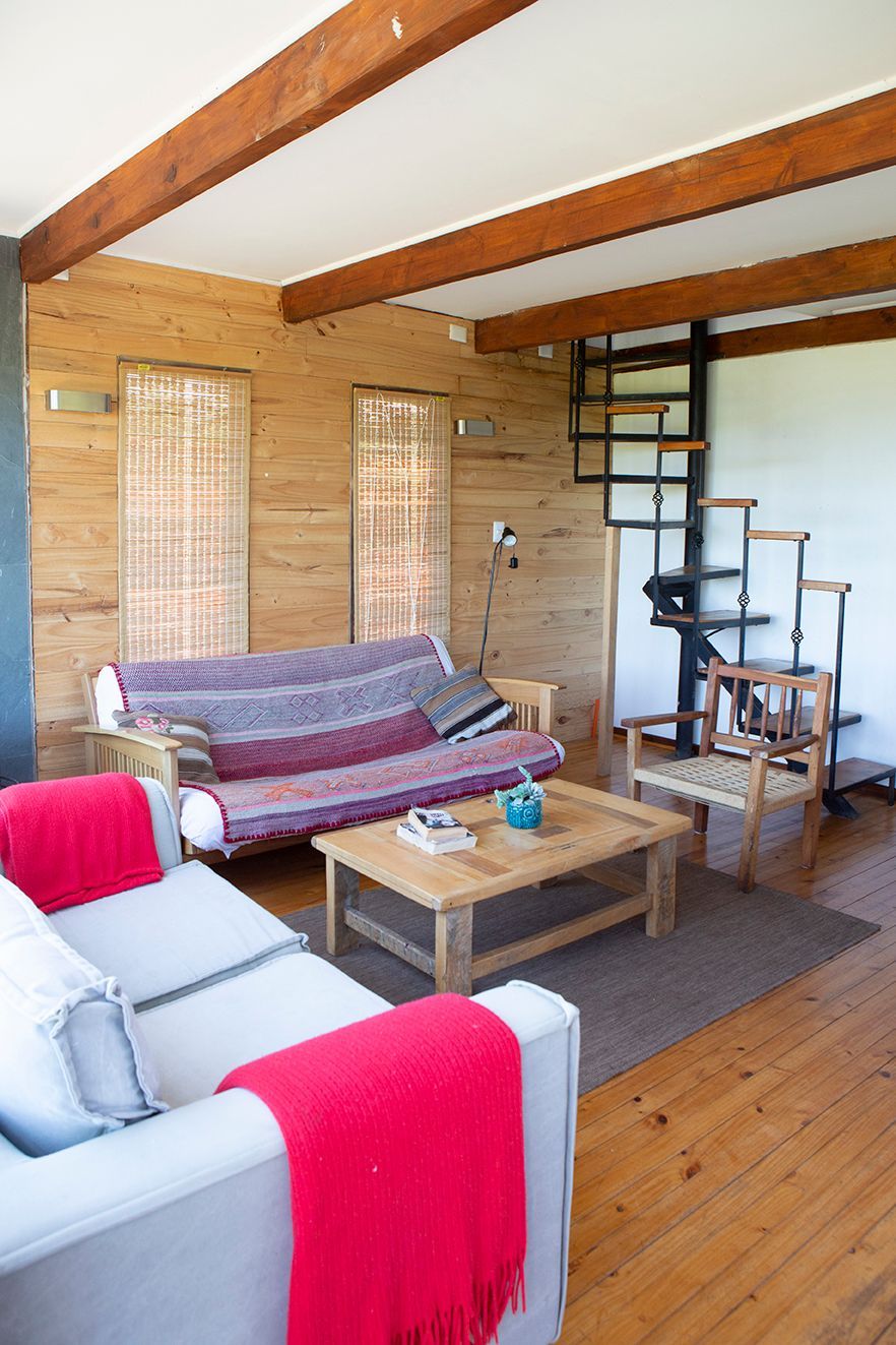 Sala de estar con vigas de madera, sofá gris claro con manta roja, futón de madera y escalera de caracol.
