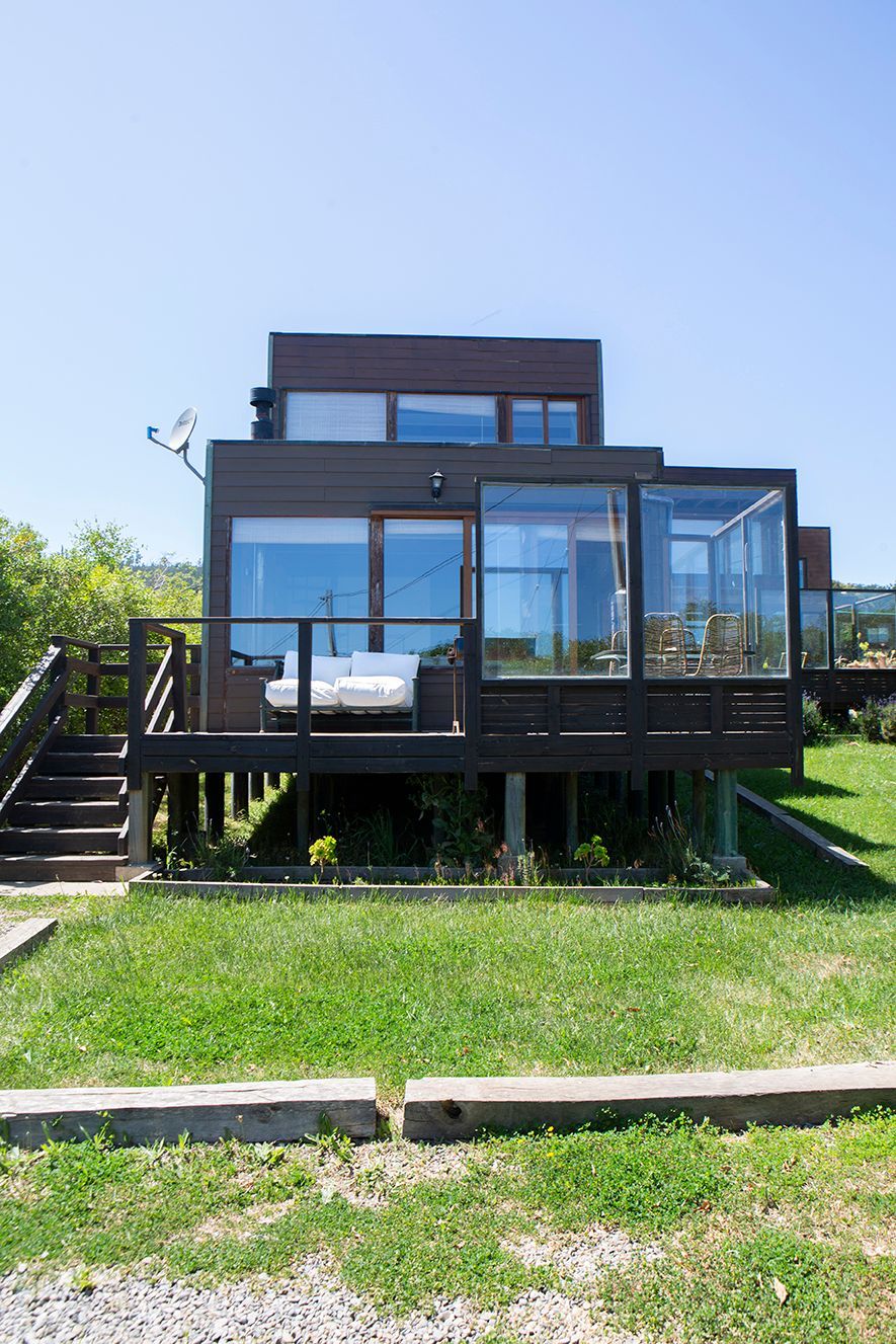 Moderna casa de madera marrón con terraza, grandes ventanales y porche cerrado con vidrio en una colina cubierta de hierba.