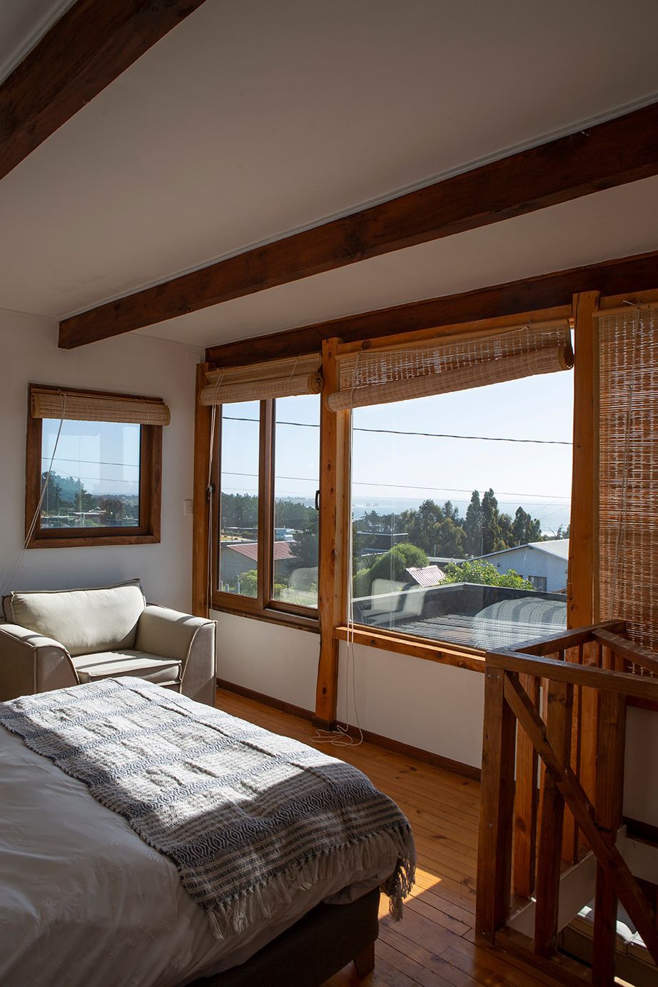 Dormitorio con vistas: cama, sillón, vigas y marcos de madera, grandes ventanales y paisaje exterior.