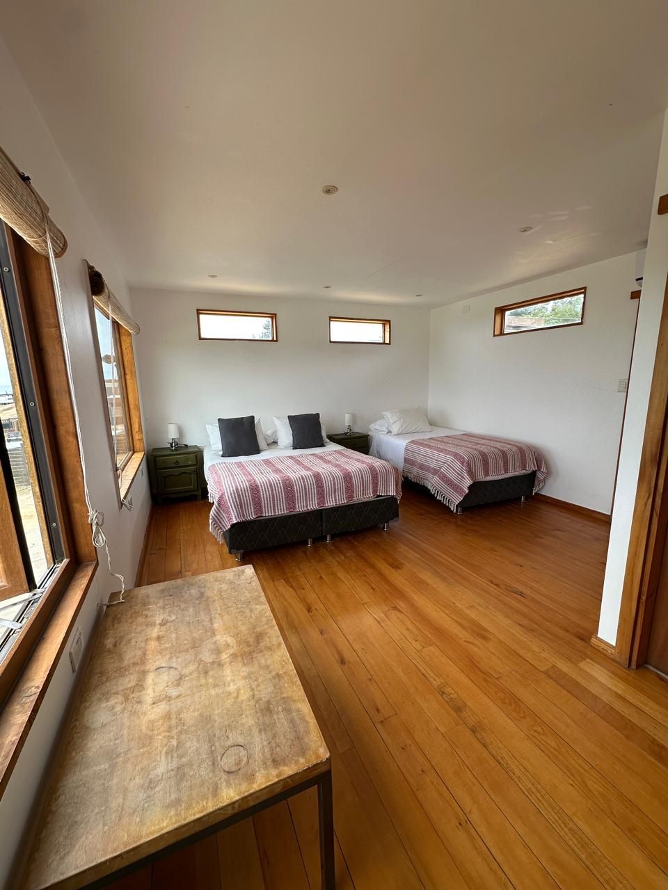 Dormitorio con piso de madera, dos camas con mantas a cuadros, mesas de noche y una mesa de madera.