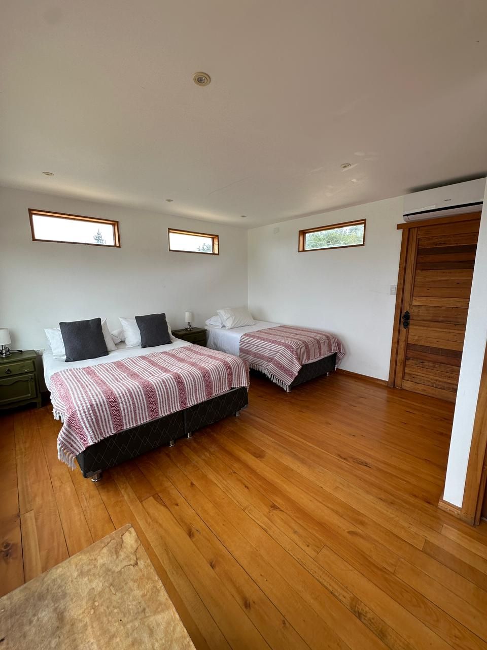 Dormitorio con dos camas, una de matrimonio y otra individual, con suelo de madera y paredes blancas.