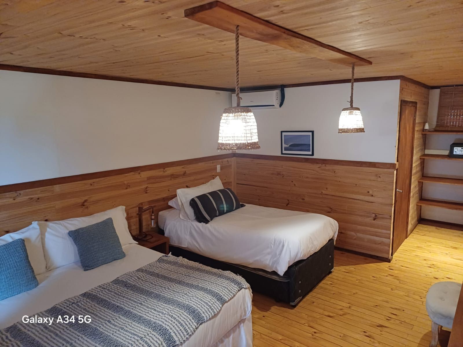 Dormitorio con dos camas, paneles de madera, lámparas colgantes y un pequeño cuadro en la pared.