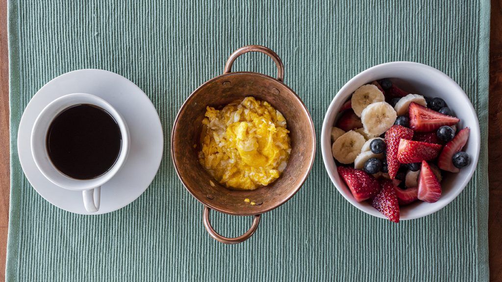 Desayuno: café, huevos revueltos en sartén de cobre, ensalada de frutas (fresas, arándanos, plátano) sobre un mantel verde.