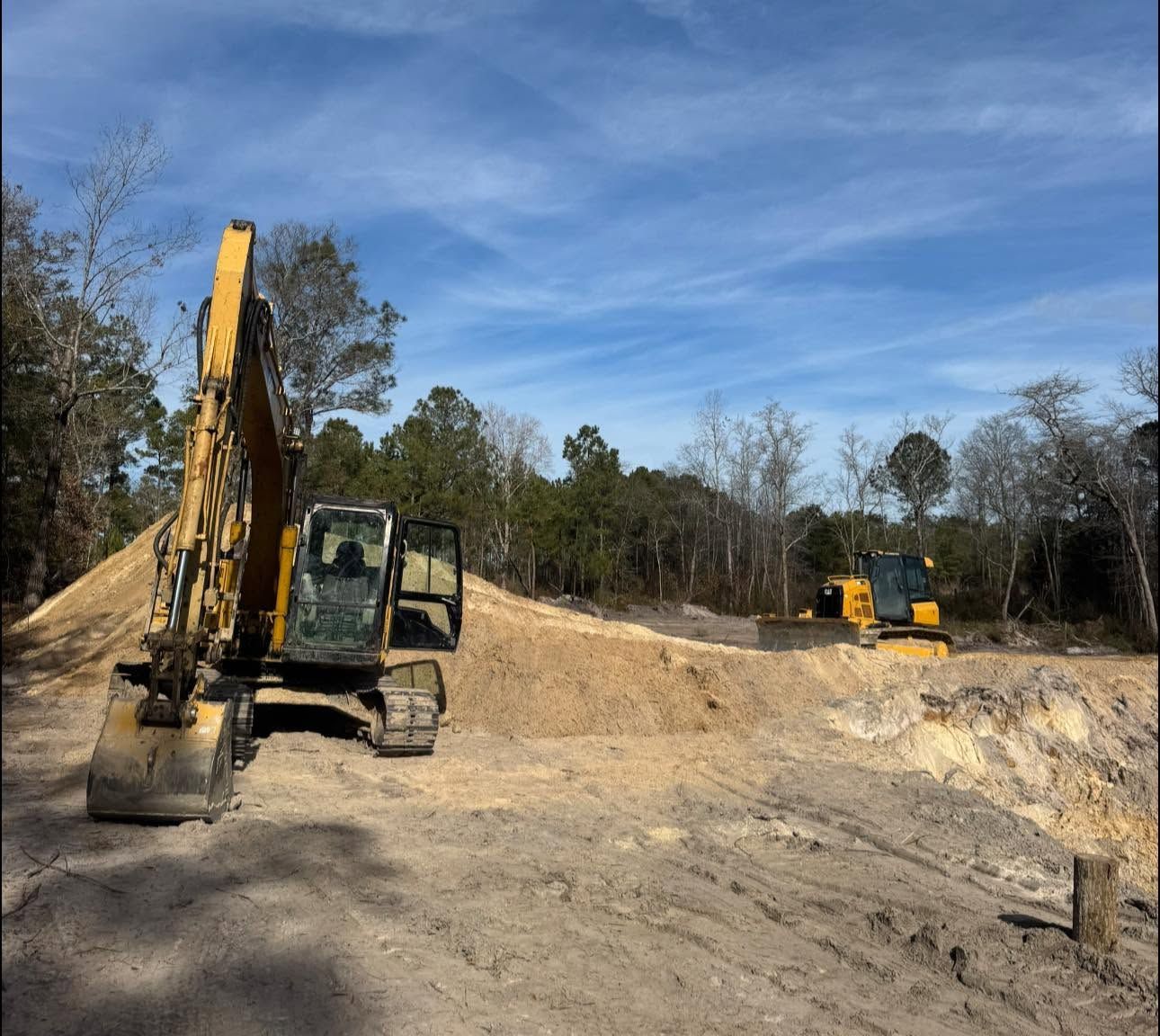 Excavators at a dirt construction site under a blue sky, with one machine in the foreground and another in the distance