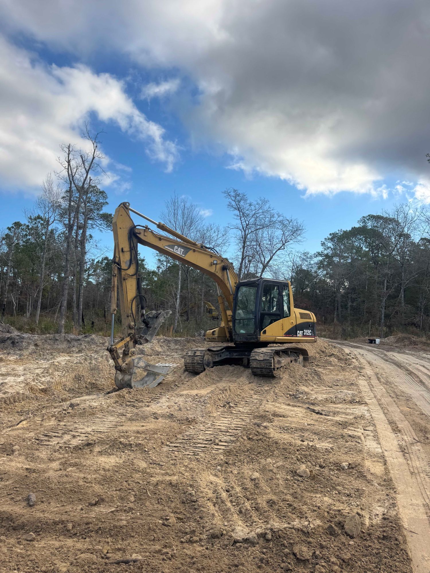 Yellow excavator digging on a muddy clearing beside a dirt road under a cloudy sky.