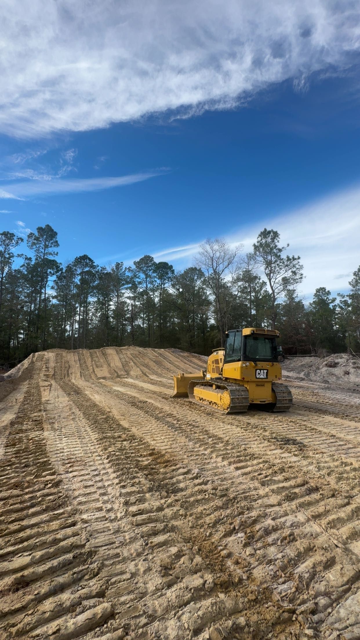 Yellow bulldozer grading a dirt road in a wooded clearing under a blue sky