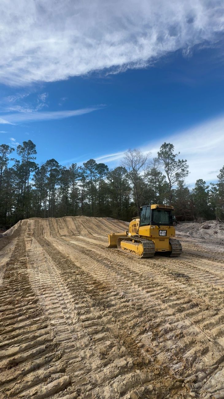 Yellow bulldozer grading a dirt road in a wooded clearing under a blue sky
