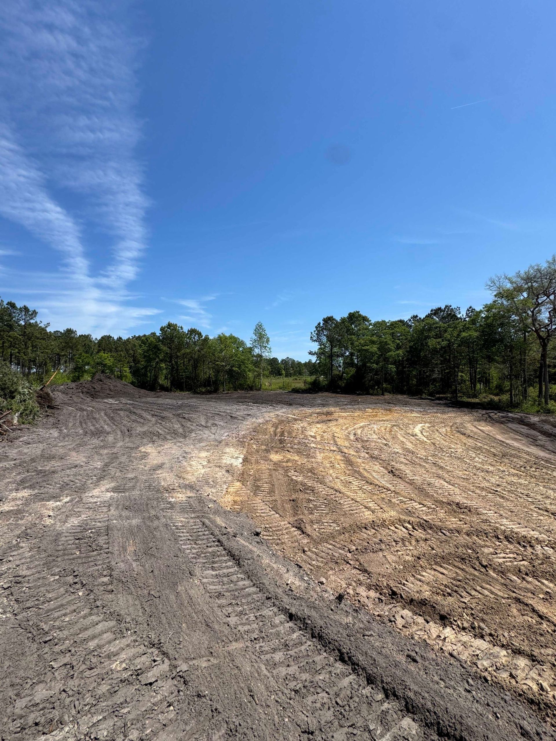 Cleared dirt lot with tire tracks under a bright blue sky, bordered by trees