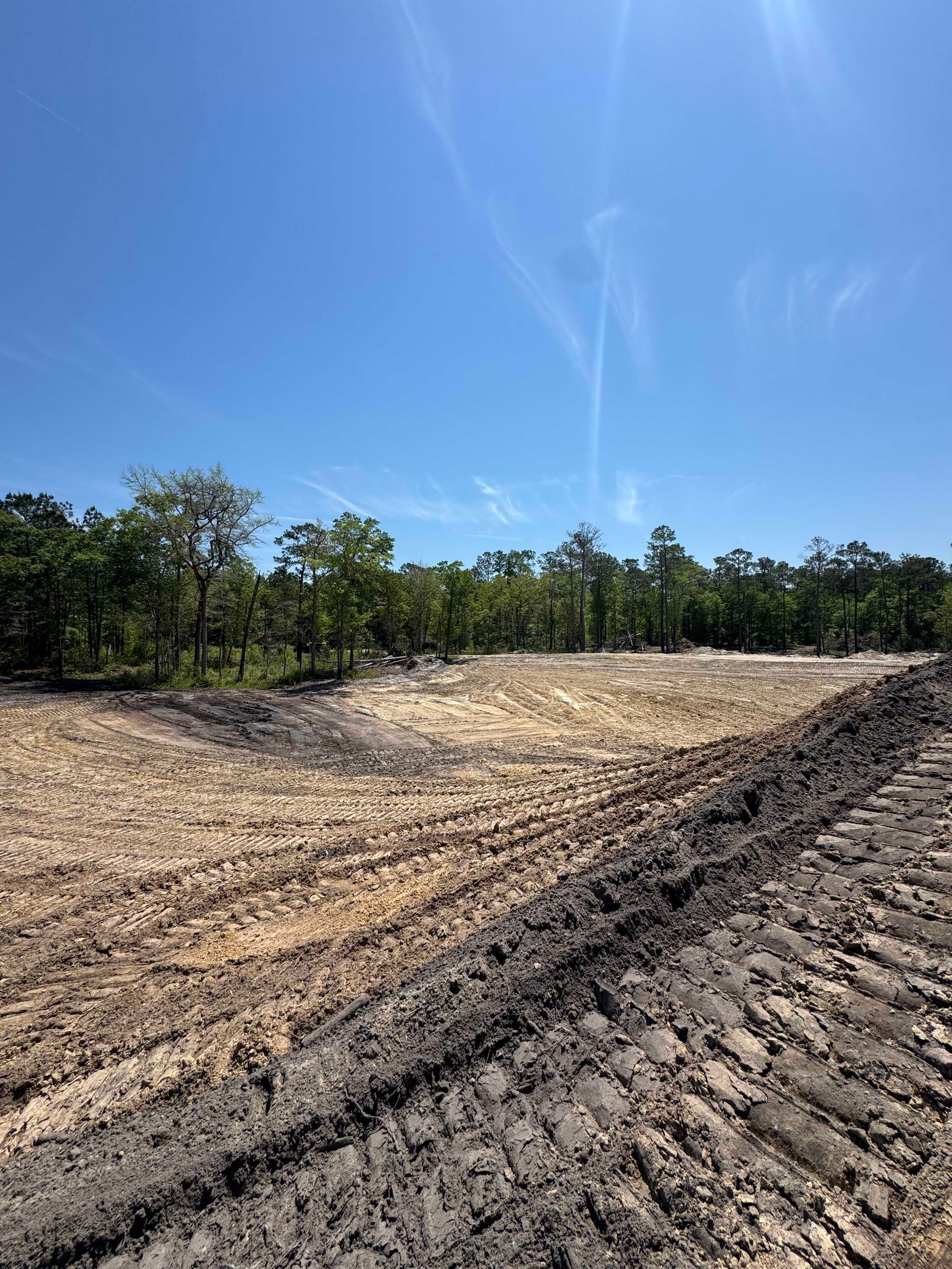 Cleared dirt construction site with tire tracks and a tree line under a clear blue sky