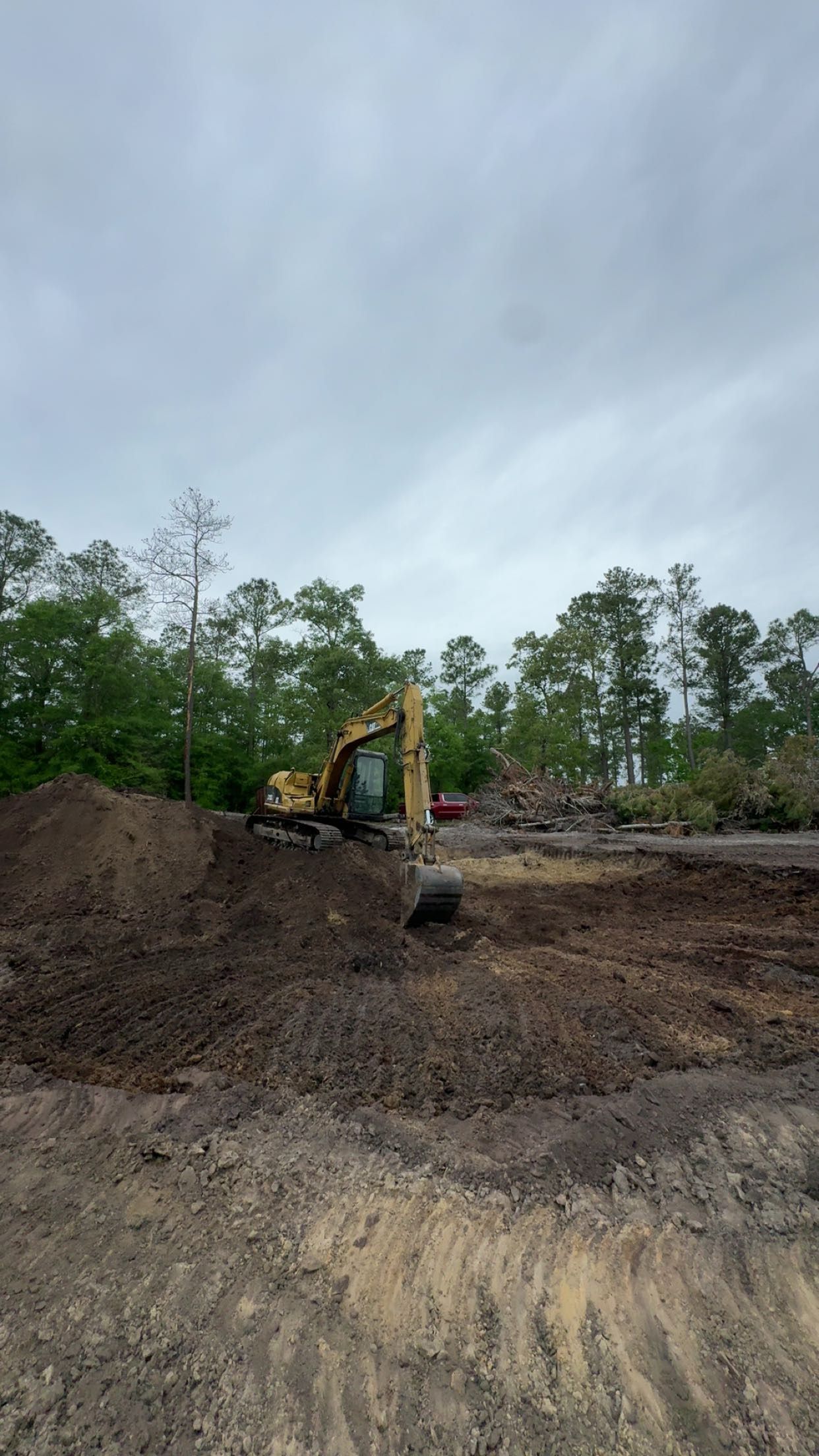 Yellow excavator on a muddy construction site under a cloudy sky, with dirt piles and trees nearby