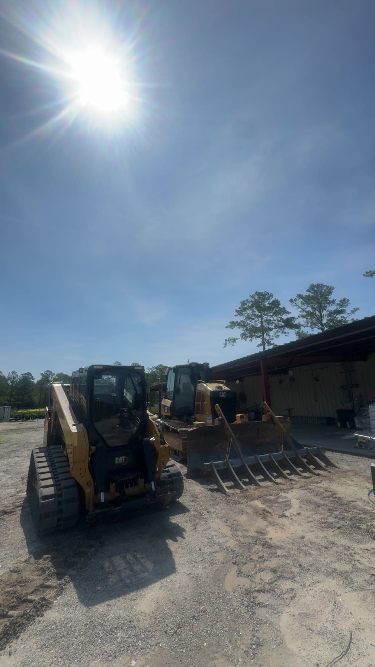 Construction equipment parked on a gravel lot beside a building under a bright sun