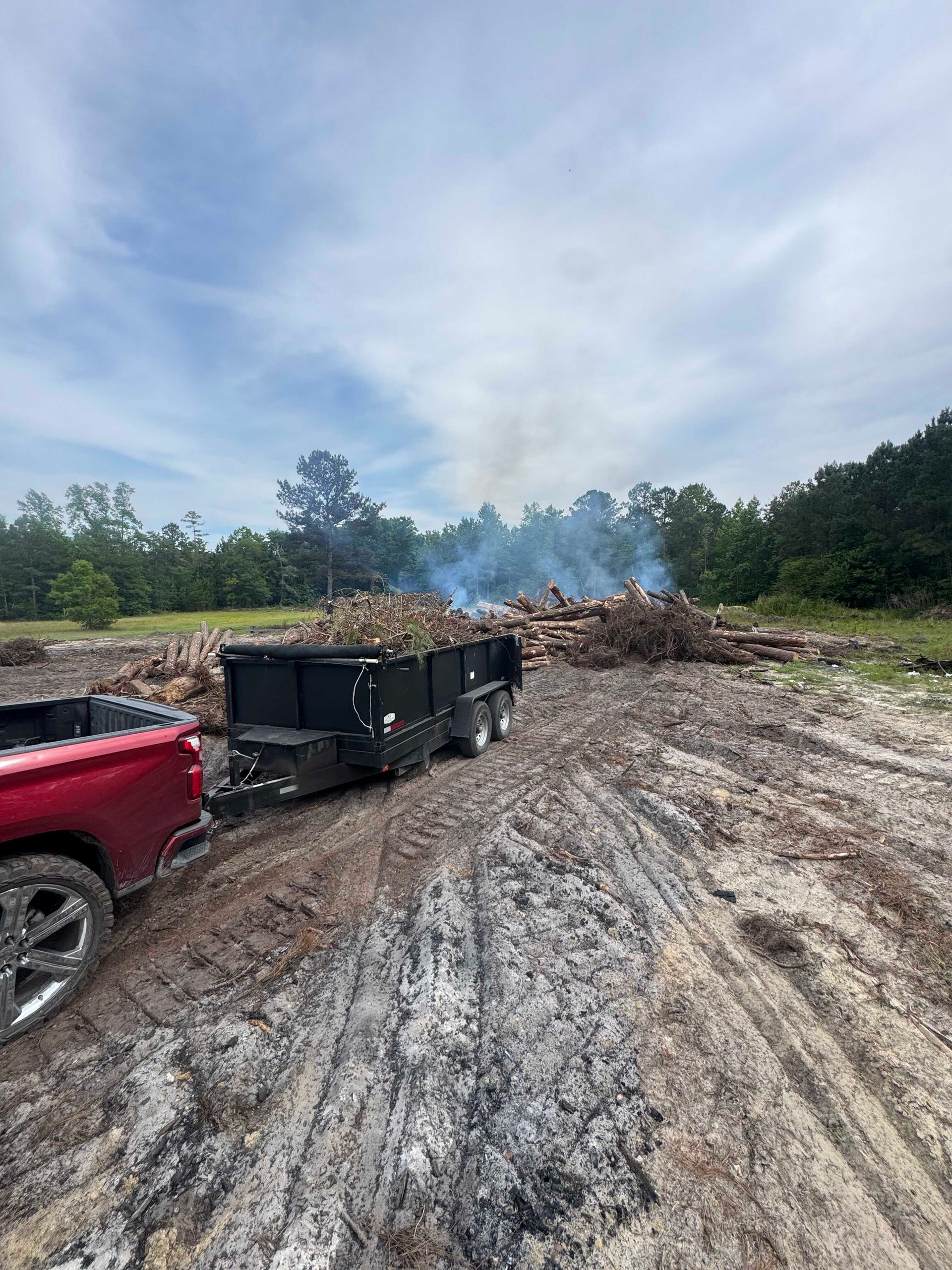 Burned debris pile in a muddy clearing with a smoking dump trailer and red truck under a cloudy sky