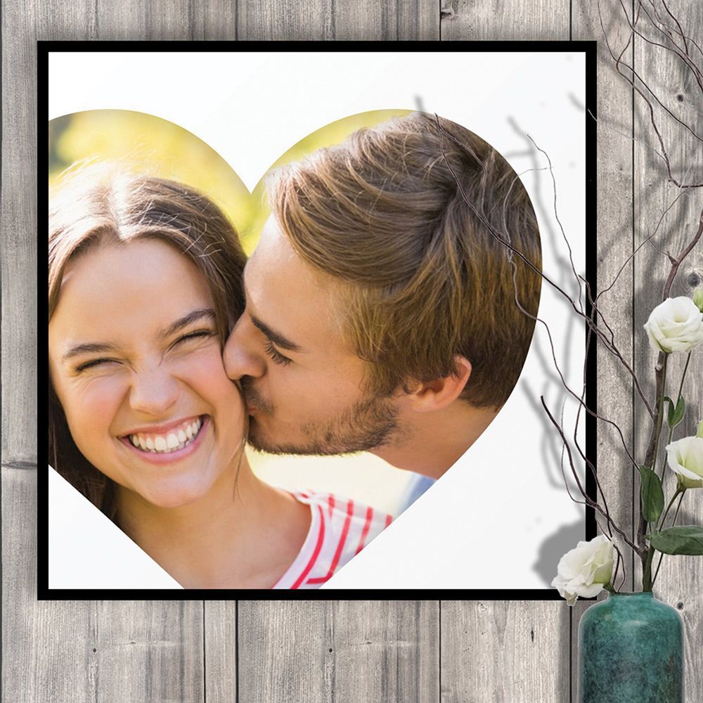 Handing collage cropped in the shape of a heart, framed in a photo frame, hanging on a wooden panelled  wall, with a vase of flowers next to it