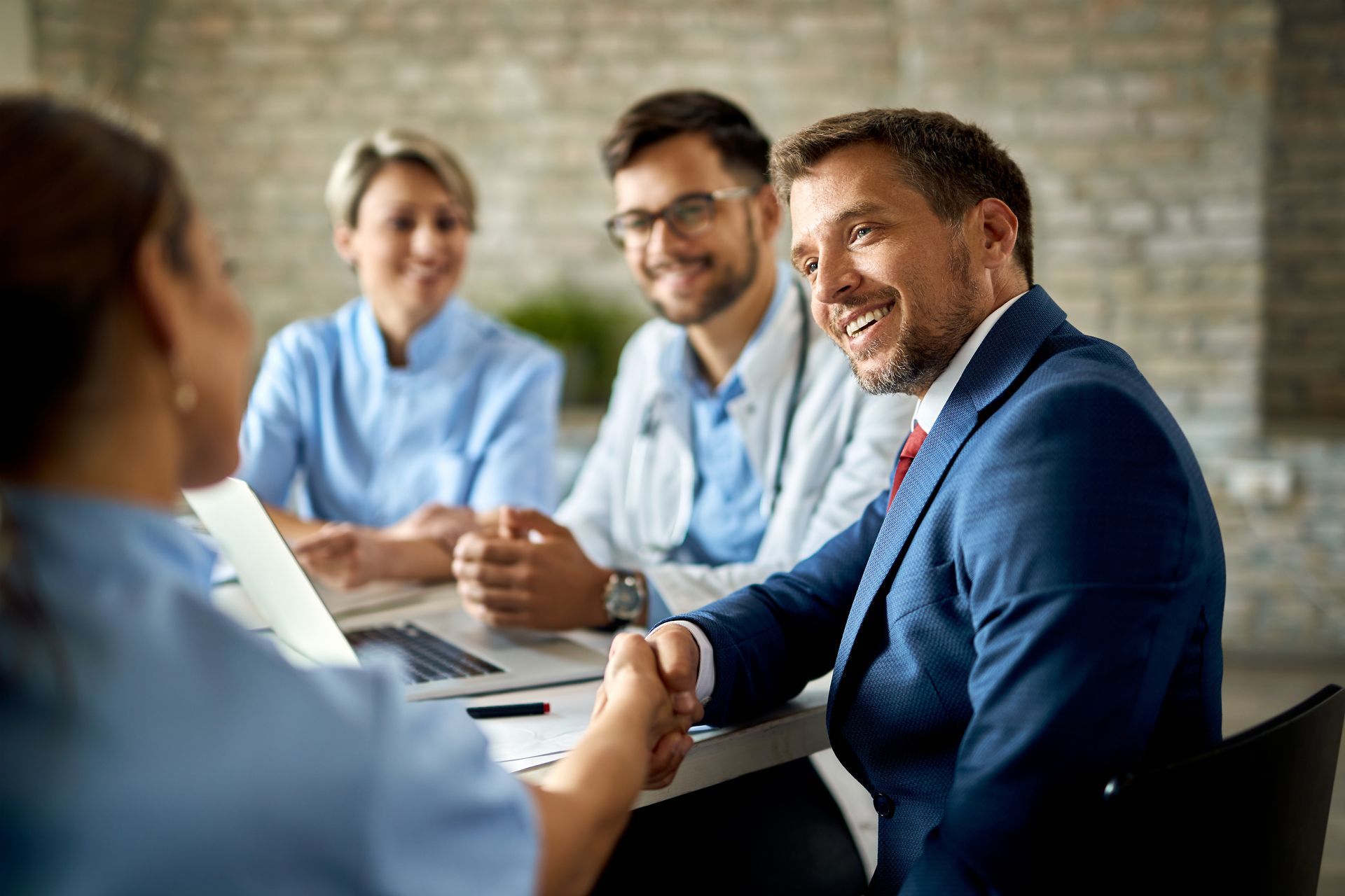 Man in suit shakes hands with person in scrubs at a table with two medical professionals.