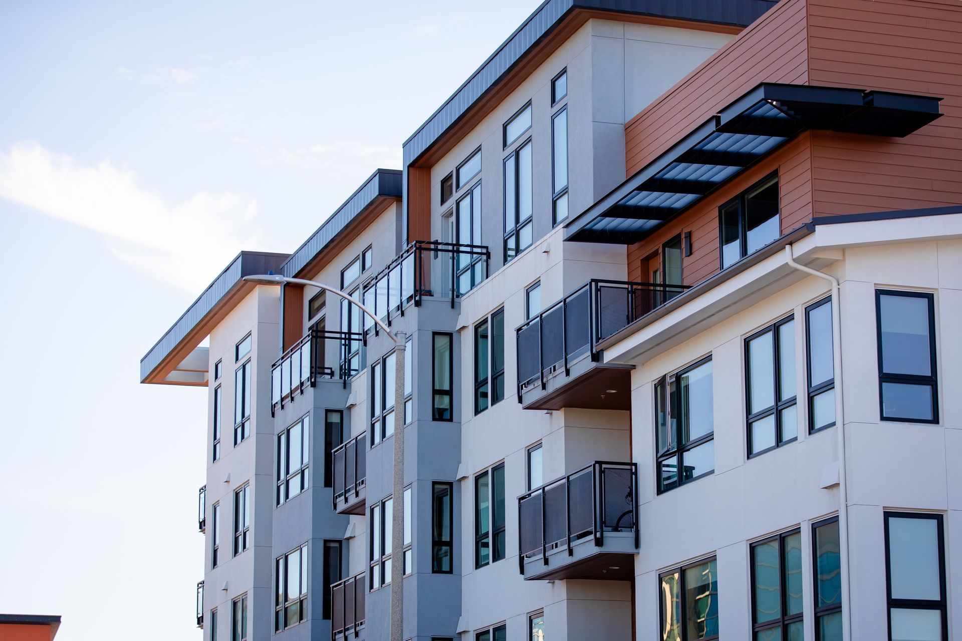Multi-story apartment building with white and brown exterior, balconies, and large windows against a blue sky.
