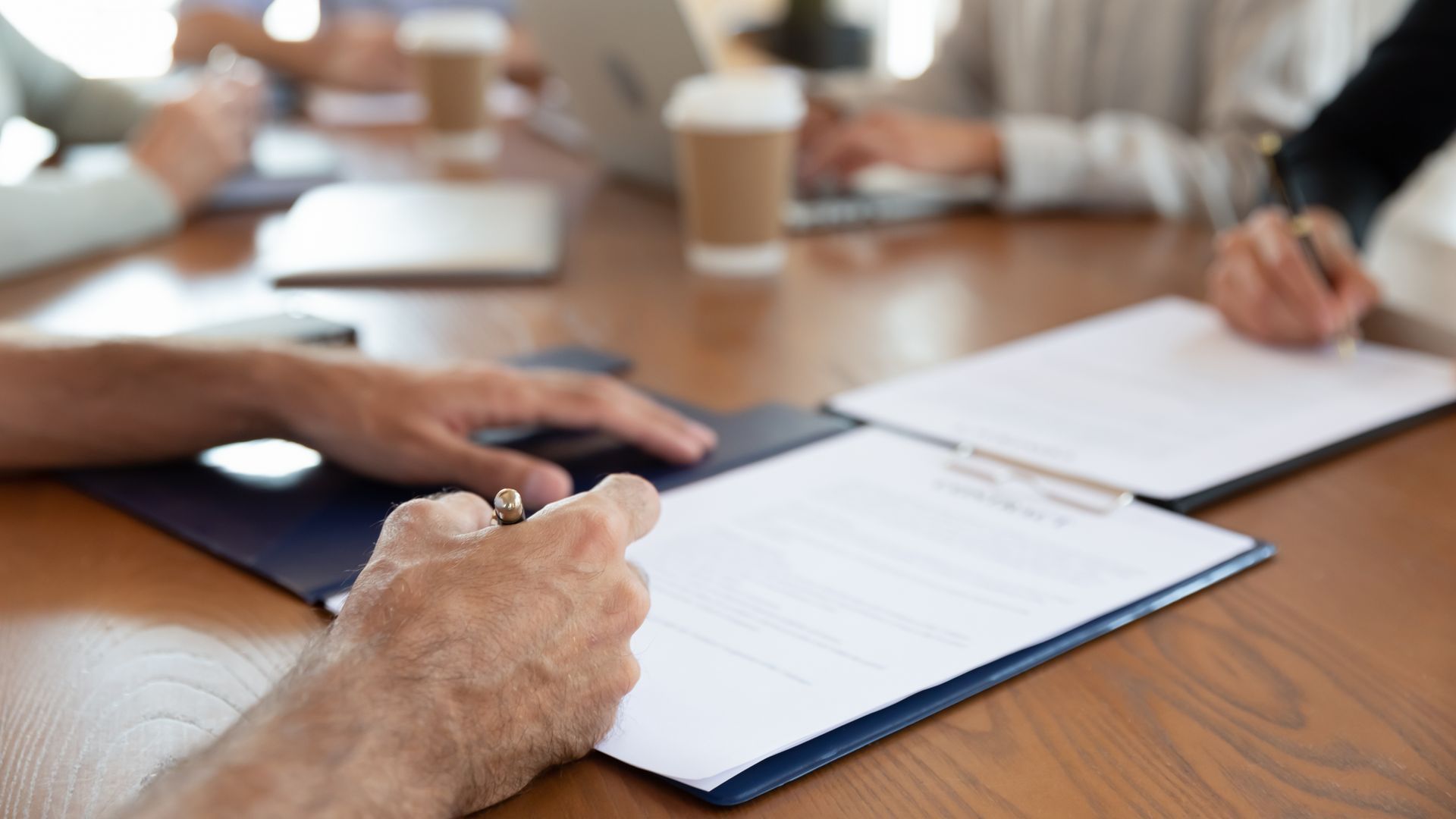 People signing documents at a conference table, other hands and laptops visible.