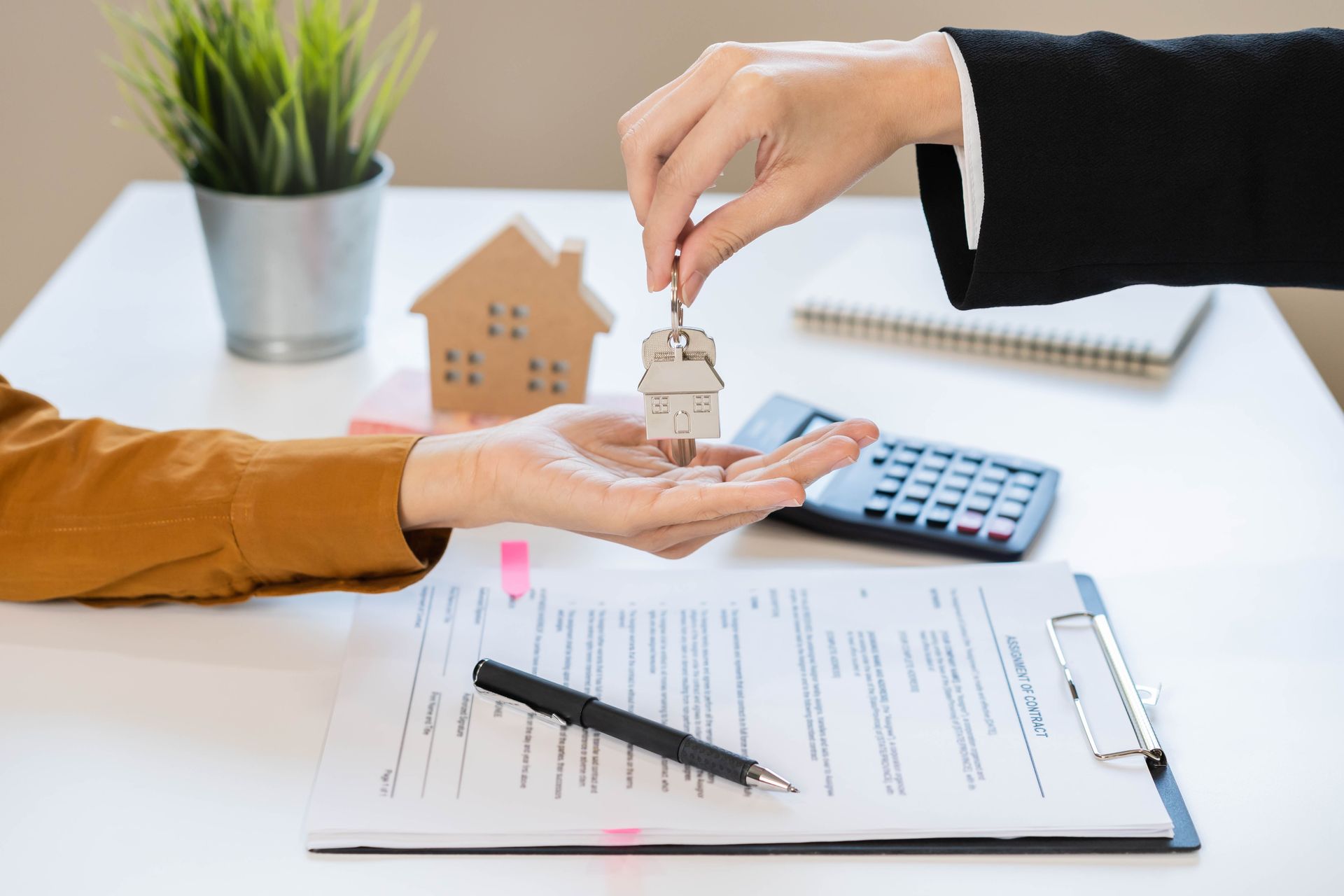 Person handing keys to another person over a document. A small house model is also on the table.