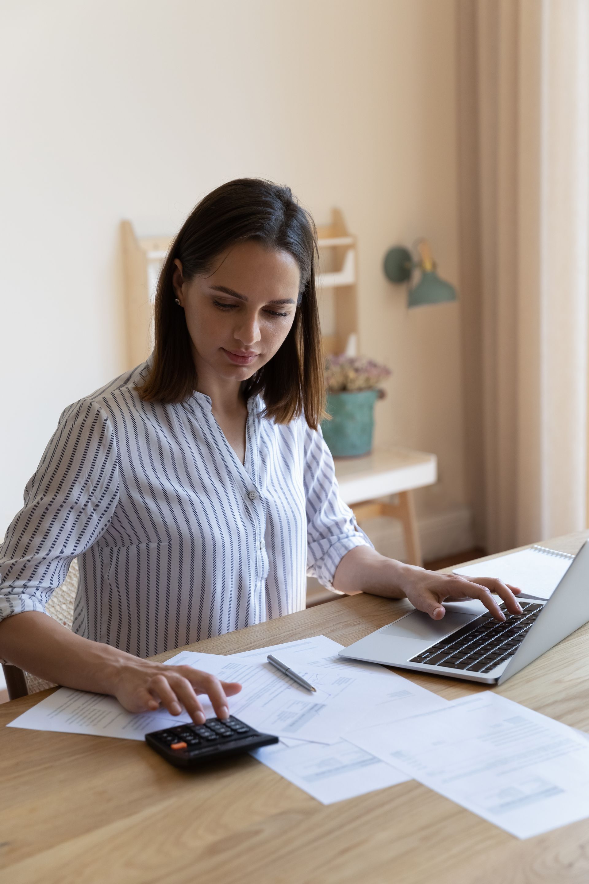 Woman at a table with papers, calculator, and laptop, focused on calculations in a home office.