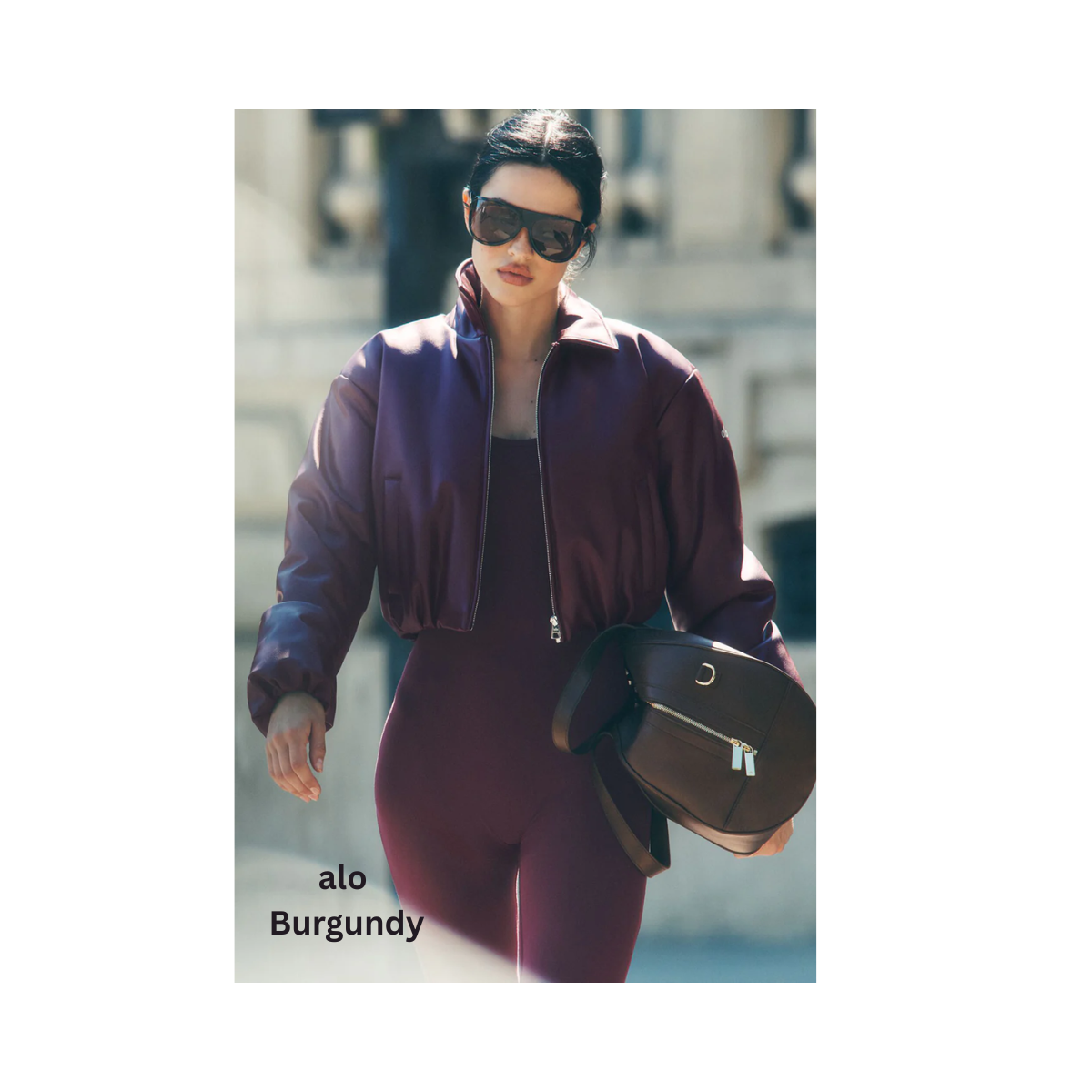 Person in burgundy outfit and sunglasses carrying a black bag while walking outdoors