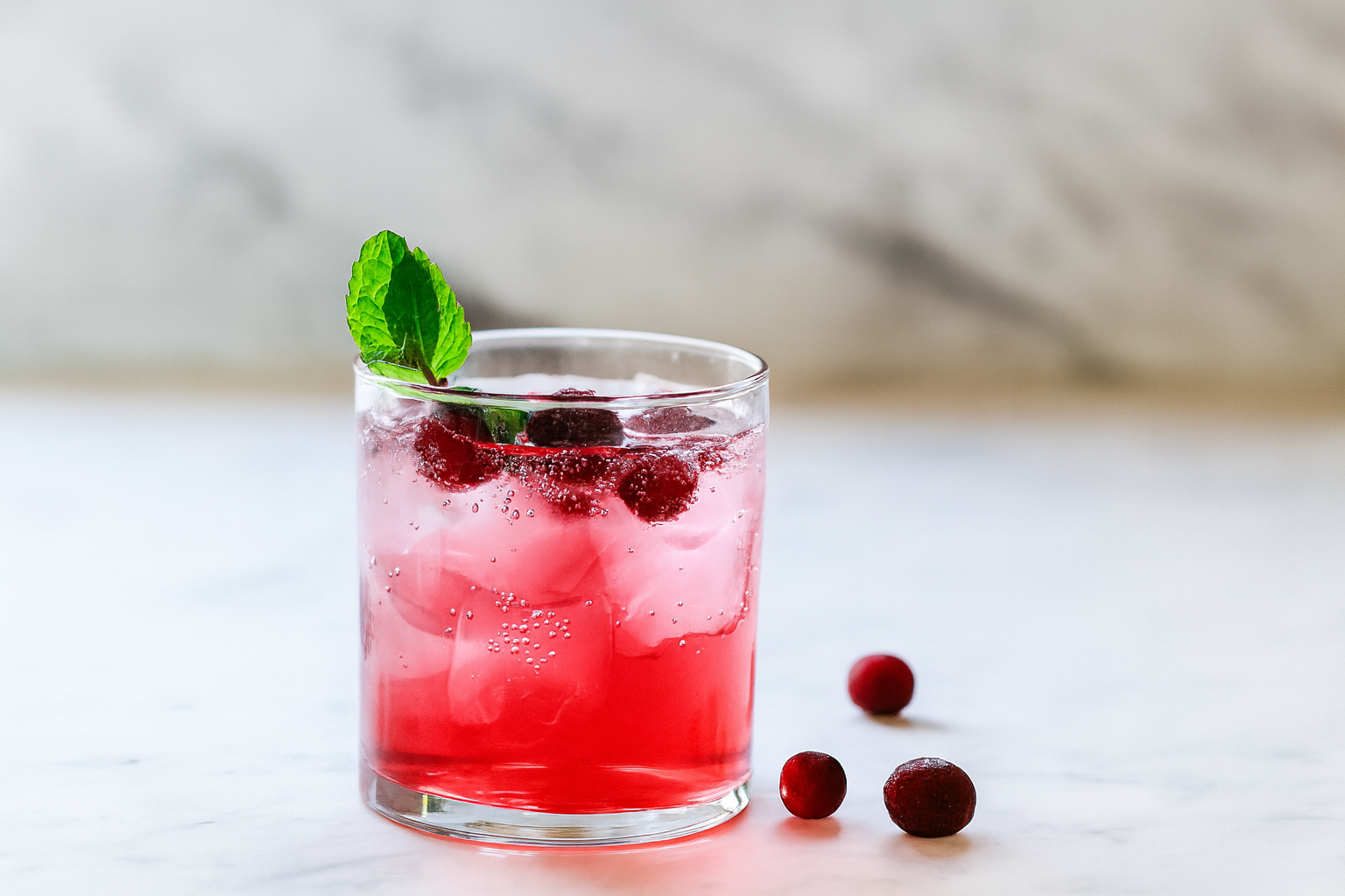 Glass of pink cocktail with cranberries, ice, and mint sprig; marble background.