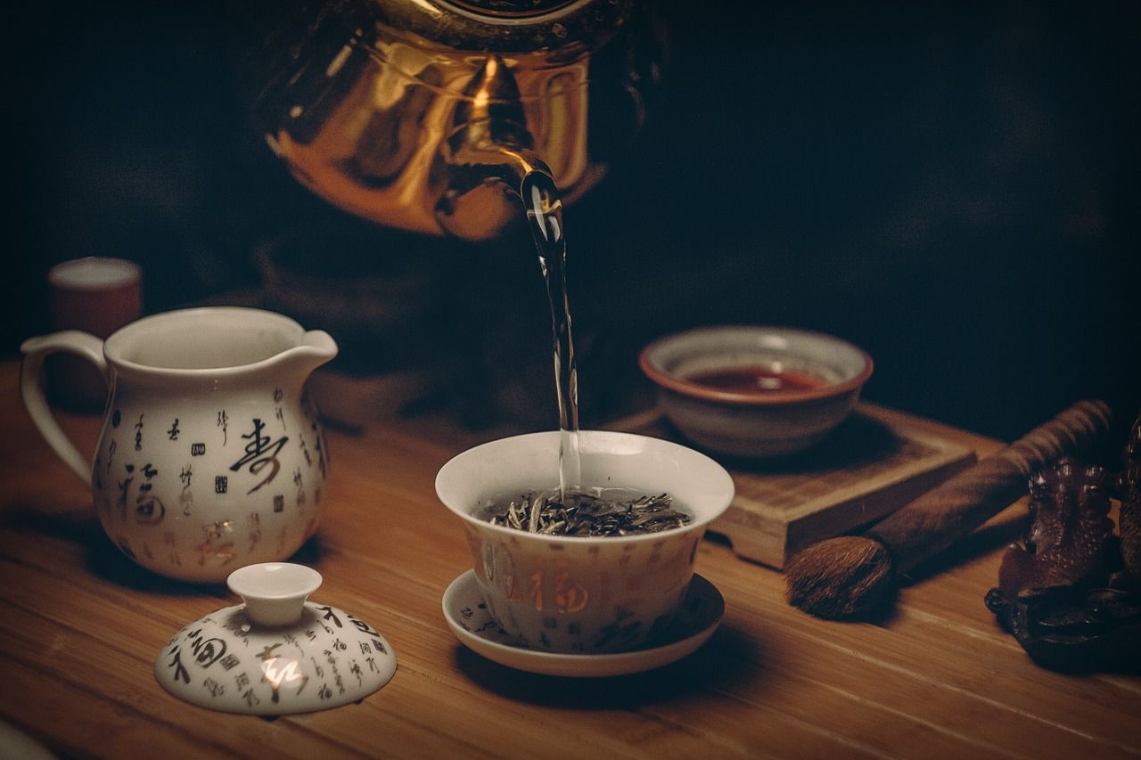 Teapot pouring tea into a cup on a wooden table with a teacup, saucer, and spoon in warm light