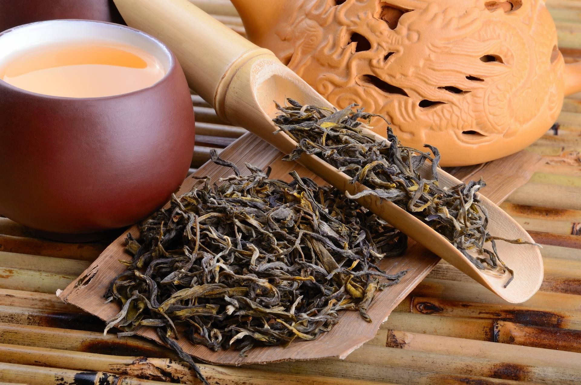 Loose tea leaves beside a cup of tea on a bamboo tray, with a wooden scoop and decorative tea set