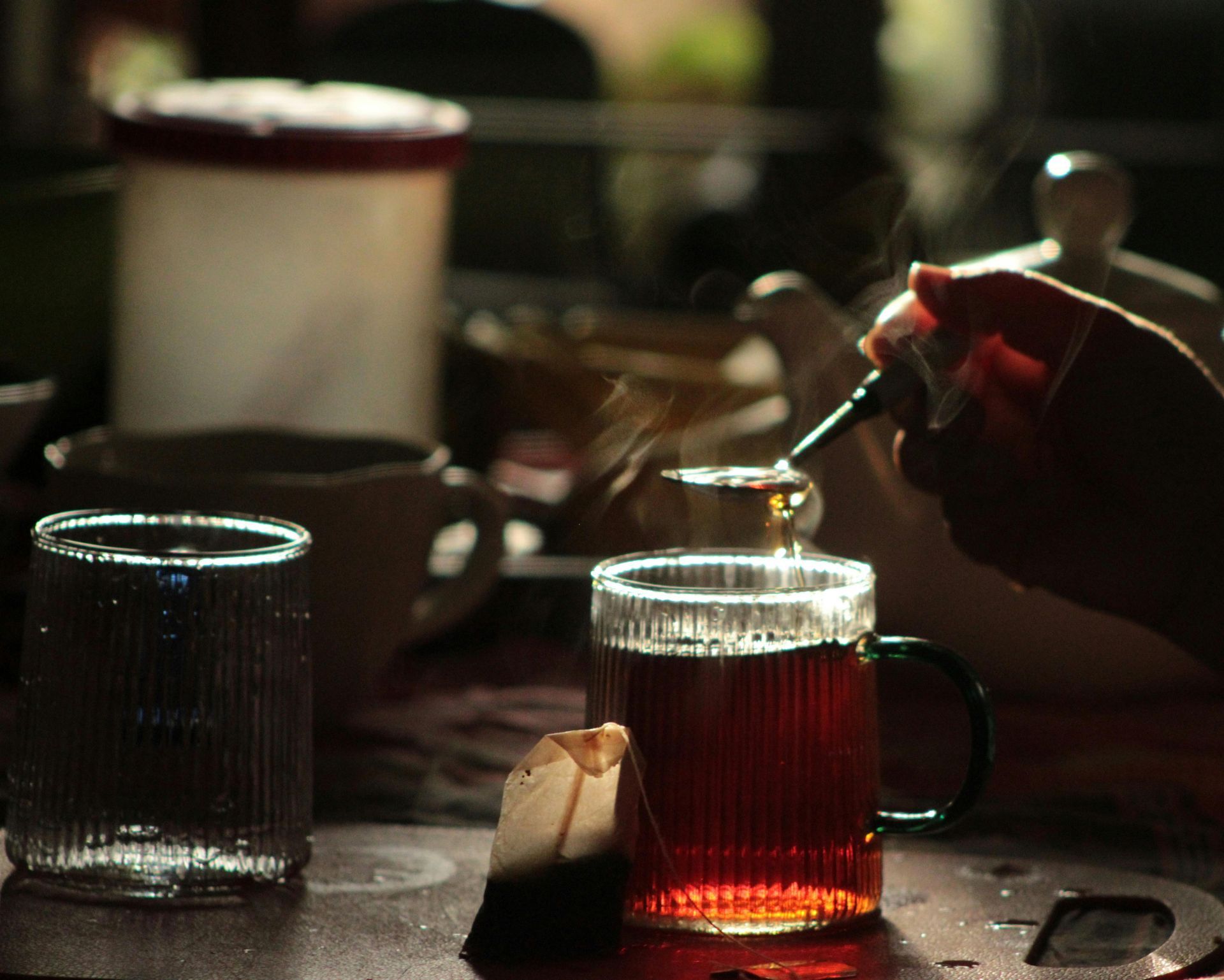 Hand pouring tea into a glass mug on a dimly lit table with other cups