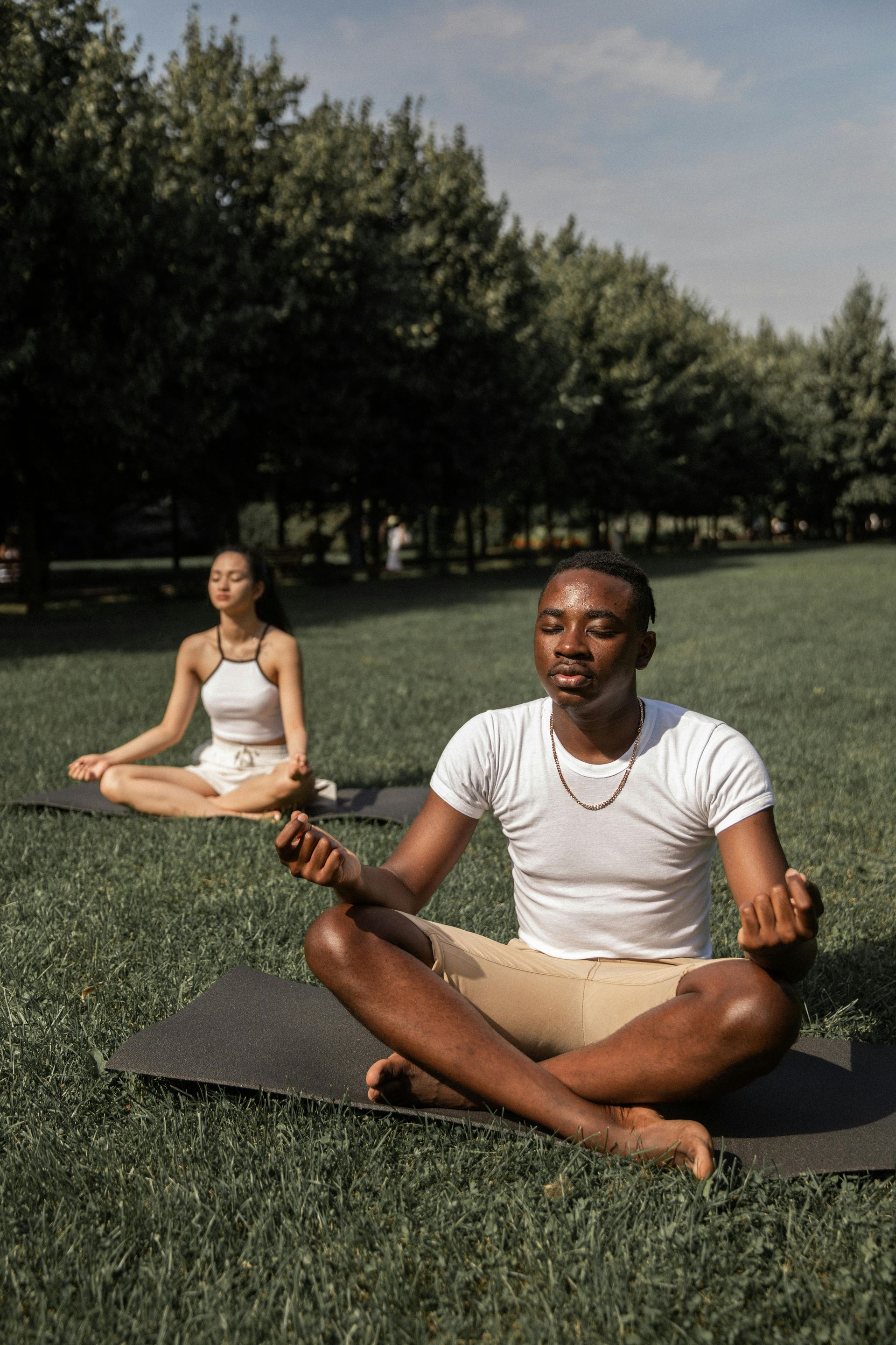 Two people meditate on yoga mats in a grassy park under trees.