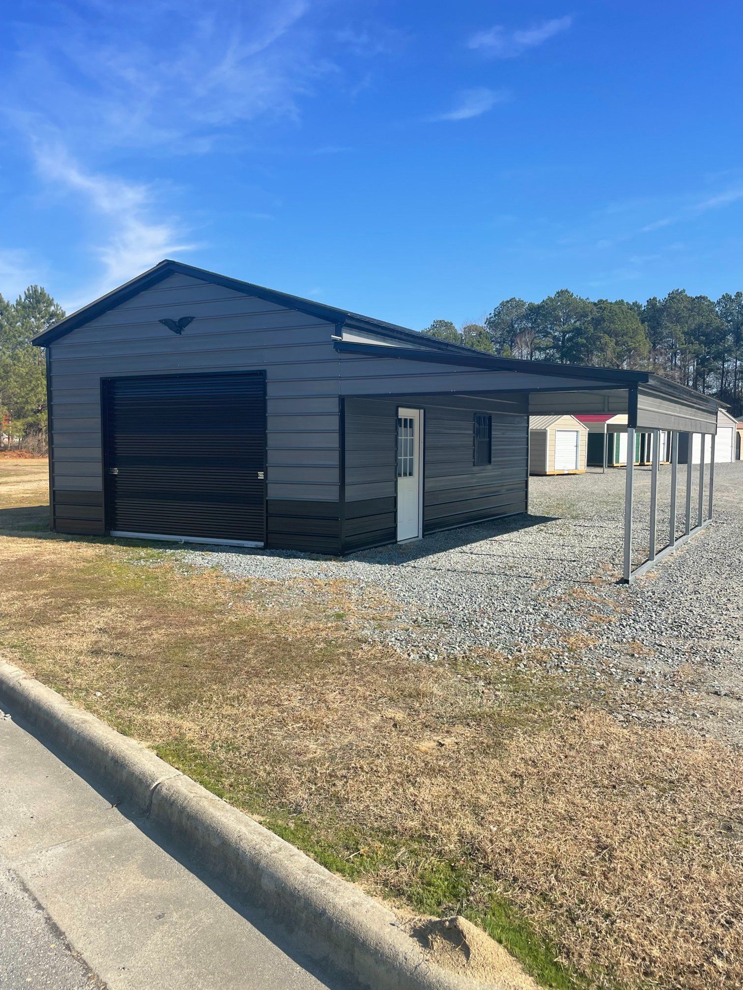 Gray and black metal storage building with a roll-up door and gravel lot.