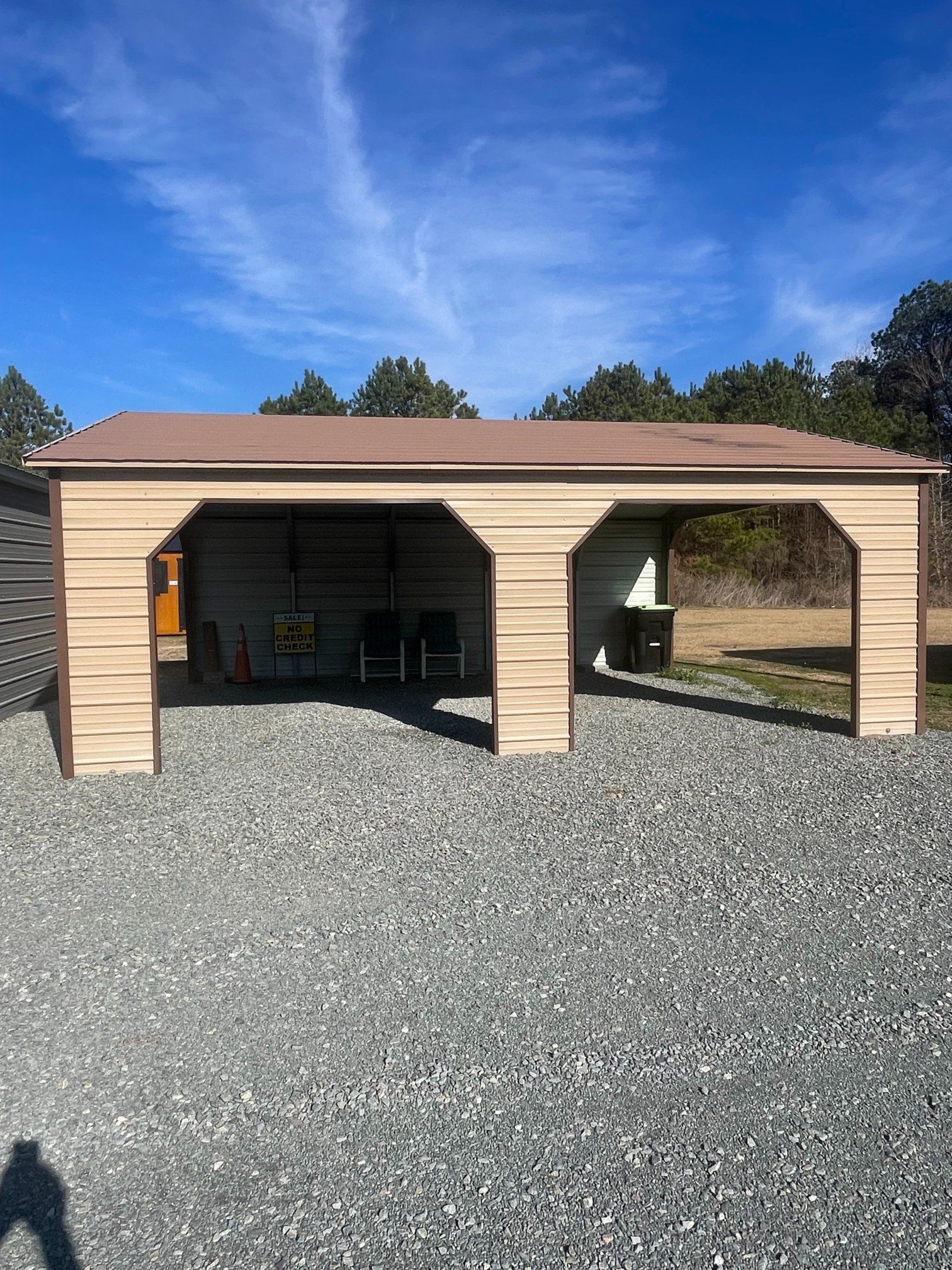 Beige metal carport with brown roof, sitting on gravel with a blue sky background.