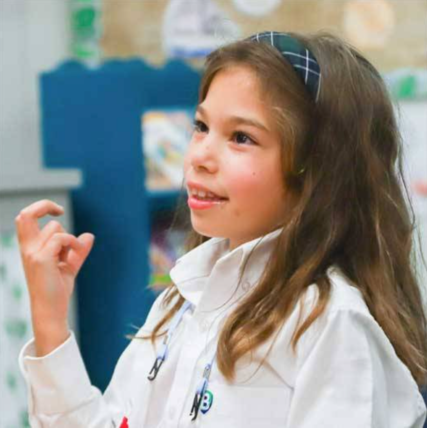 A little girl in a white button down and blue headband doing sign language in a classroom 