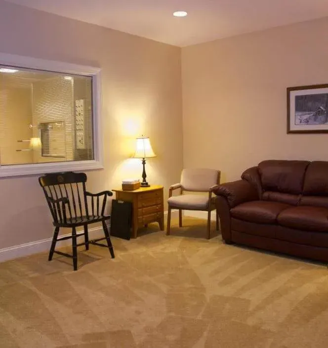 Cozy living room with brown sofa, armchair, lamp, rocking chair, and a large window into another room