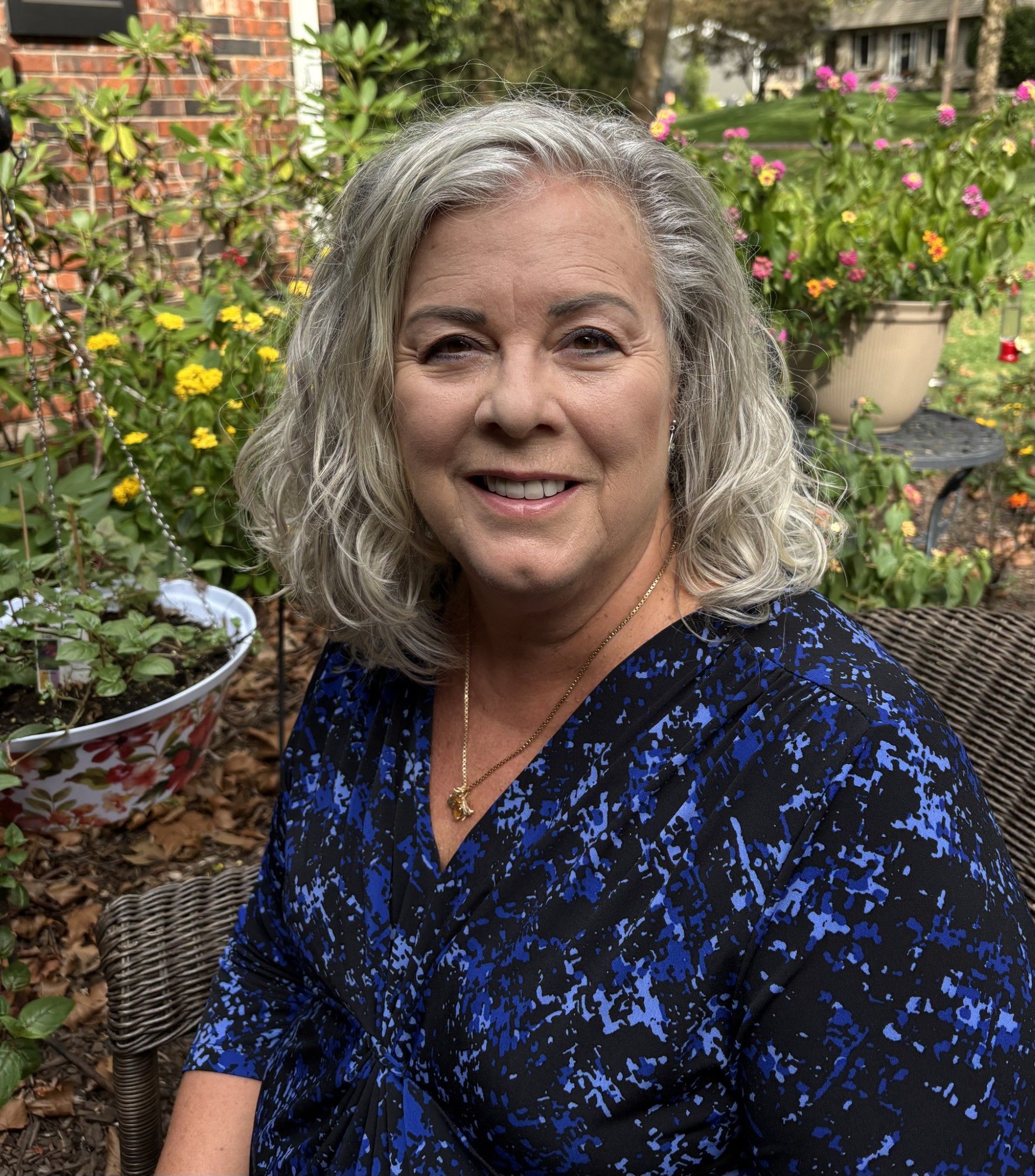 Smiling woman in a blue patterned top seated in a flower garden on a wicker chair