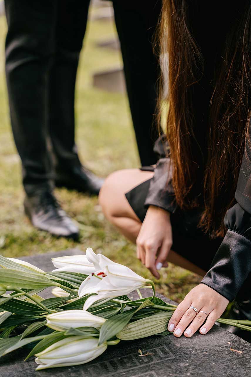 Person Mourning At A Gravesite While Placing White Lily Flowers On A Granite Headstone In A Cemetery Setting