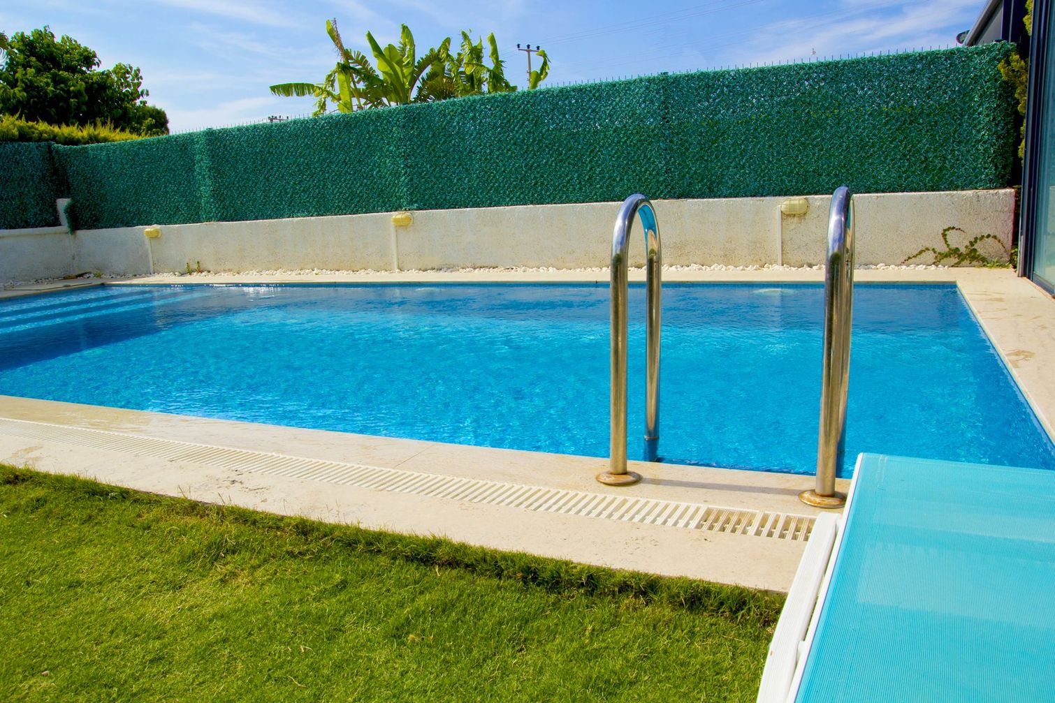 Piscina blu con scaletta in metallo, terrazza bianca e prato verde sotto un cielo soleggiato