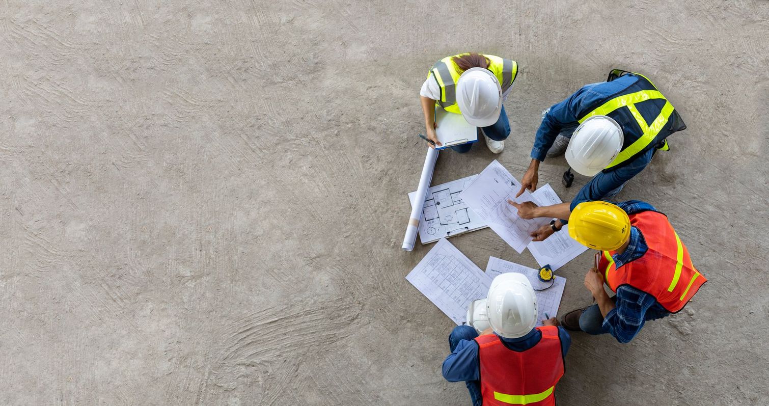 Operai edili con giubbotti e caschi di sicurezza esaminano i progetti su un pavimento di cemento.