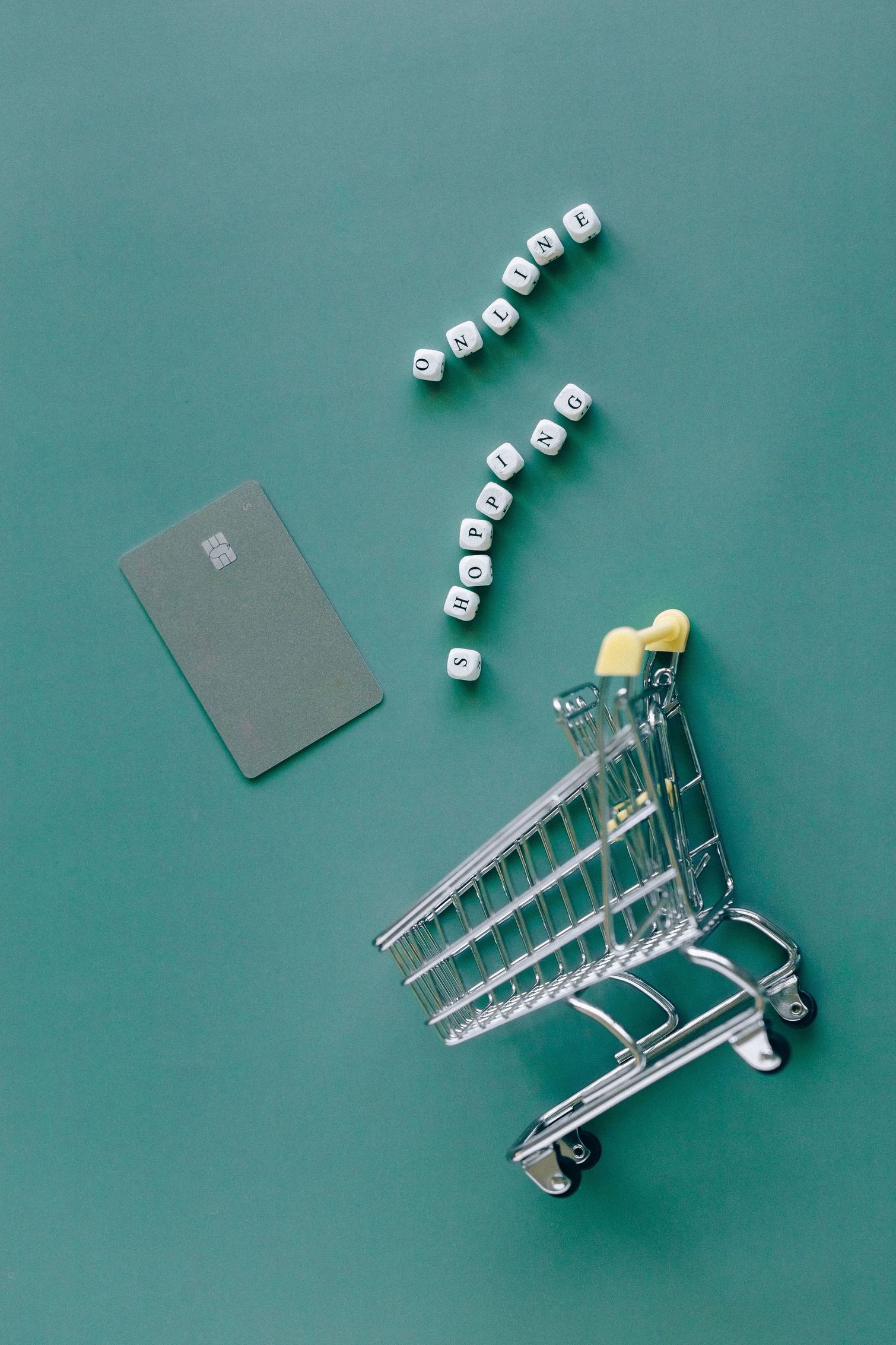 Mini carrello della spesa con oggetti gialli, cubi bianchi sparsi e un cartoncino grigio su sfondo verde acqua.