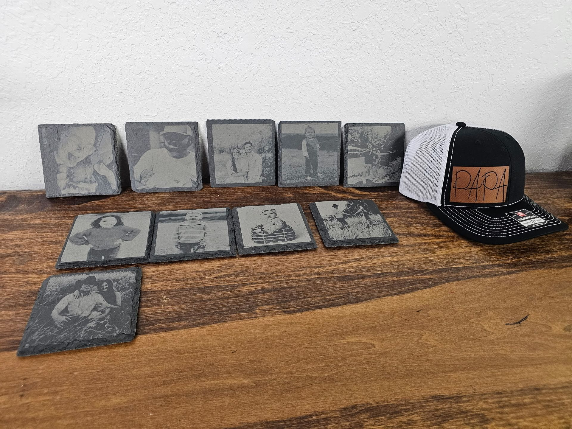 Ten square photo-engraved slates are arranged on a wooden table next to a black and white baseball cap.