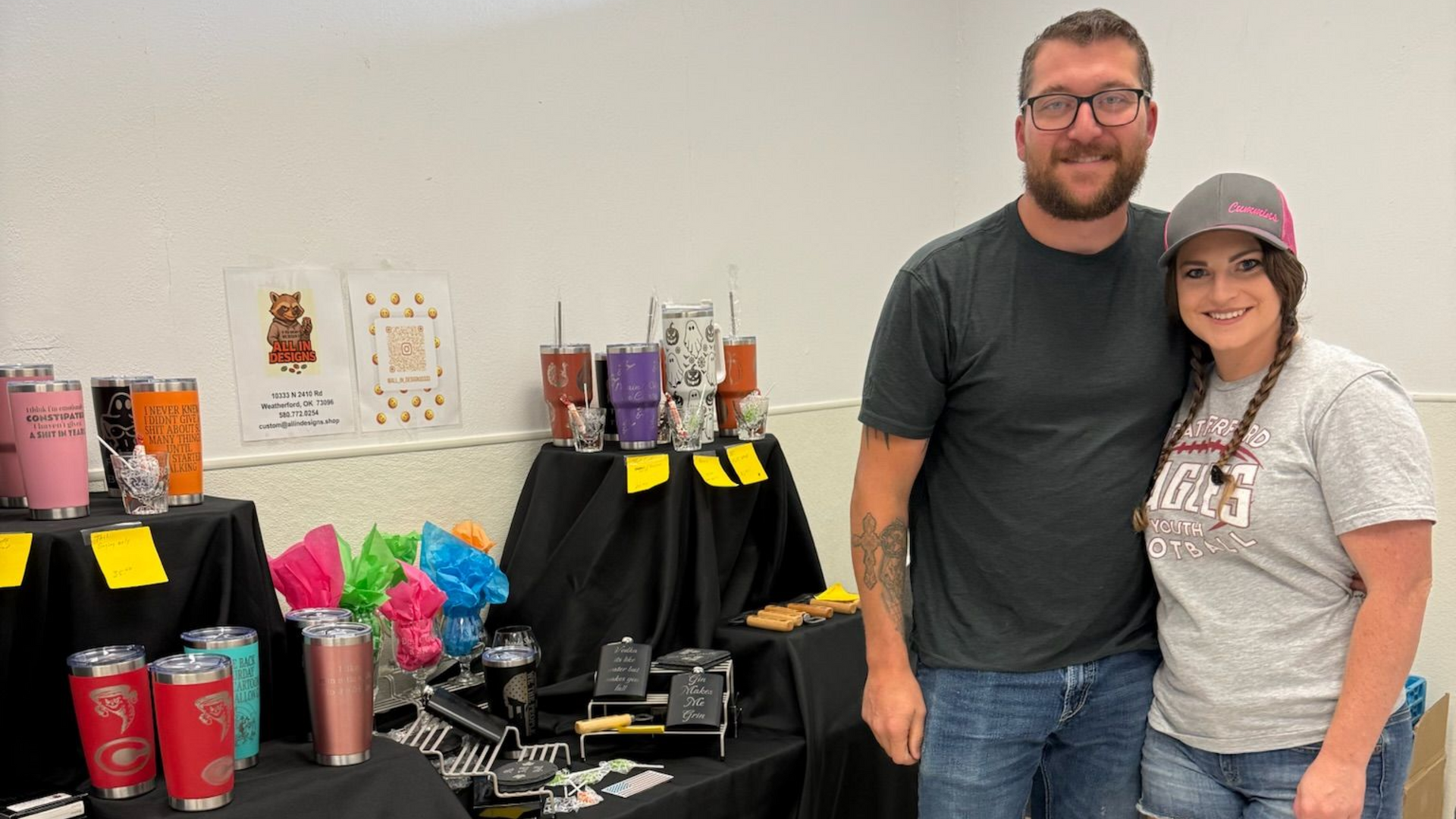 A couple stands behind a table displaying various custom-decorated tumblers and merchandise at an indoor event.
