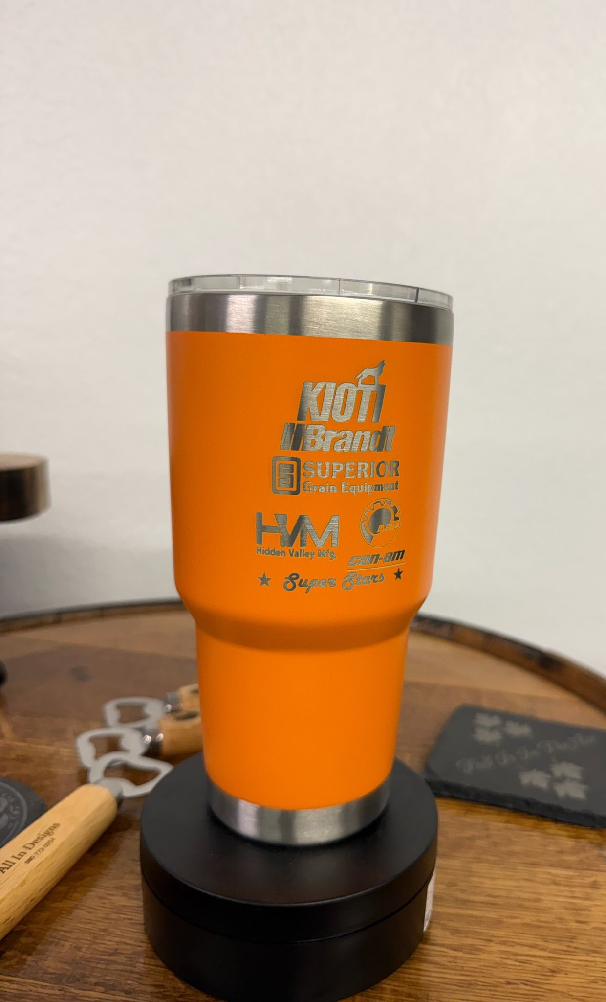 An orange stainless steel tumbler with engraved text sits on a black circular display stand on a wooden surface.