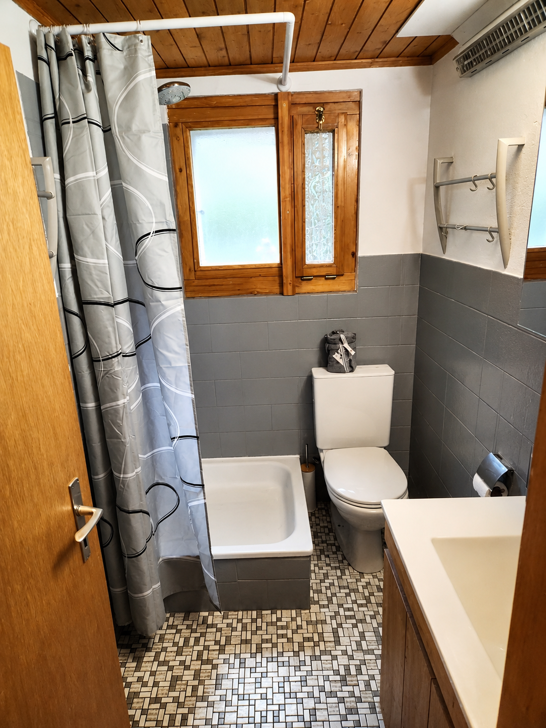 Petite salle de bains avec toilettes, rideau de douche, lavabo et fenêtre, encadrement en bois et sol carrelé à motifs.